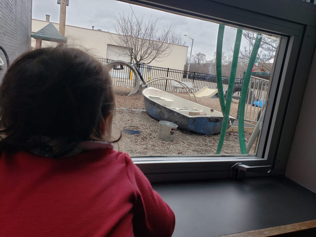 Child looks out at the playground with a boat and climbing structures.