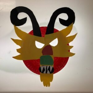 A mask of dragon featuring curly horns and a snarling face, made from a paper plate.